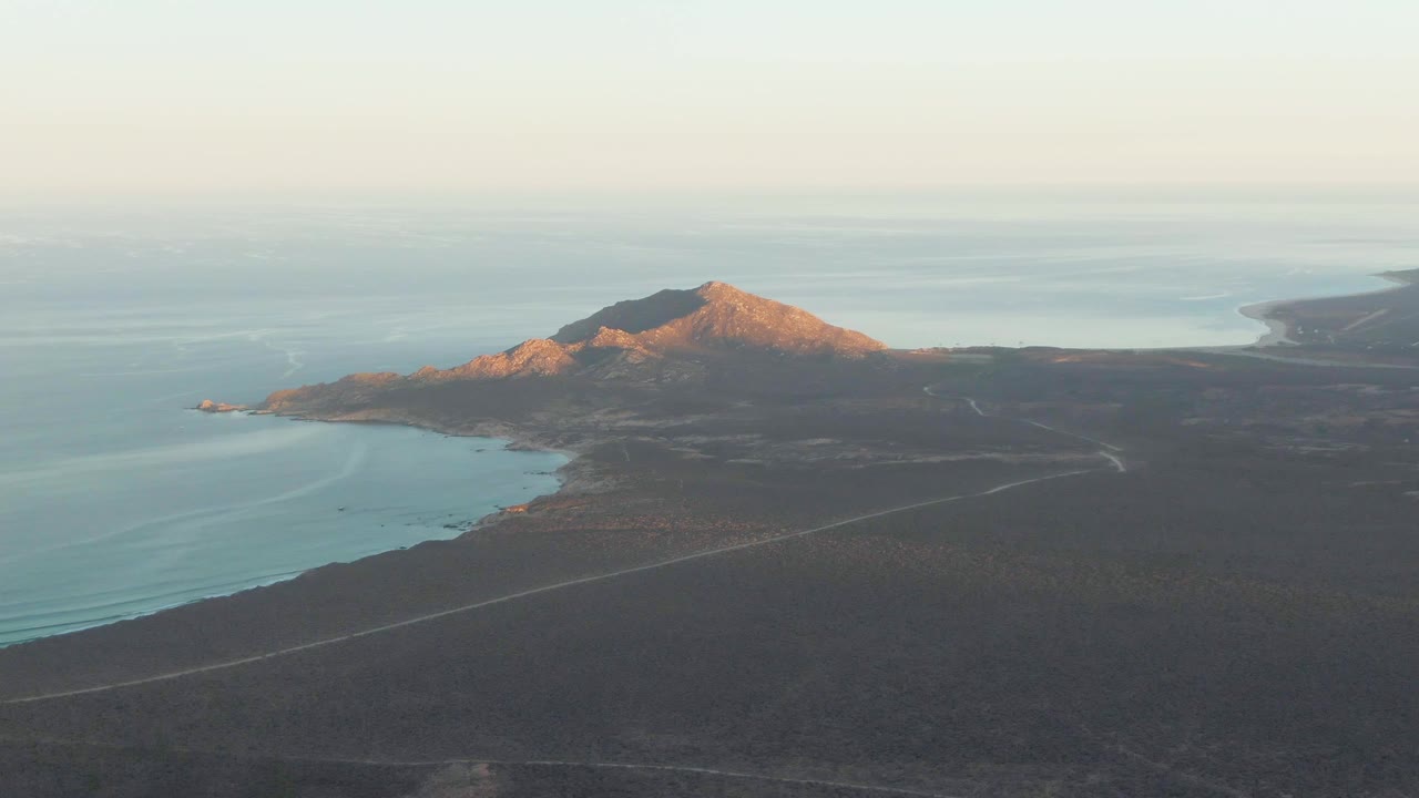 Aerial view in Cabo Pulmo with the mountain centered above dark arid land, blue water along the left shoreline, a thin road through the foreground, and no visible people.