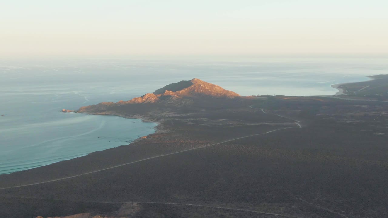 Cabo Pulmo aerial frame showing warm light on the mountain, calm ocean wrapping the coast, dark desert terrain below, a visible road line, distant haze, and no visible people.