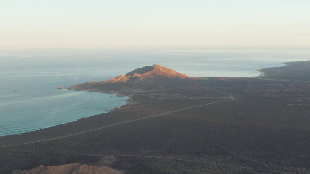 Aerial frame over Cabo Pulmo with the mountain lower in the composition, ocean on the left and beyond the ridge, dry terrain in front, faint roads, and no visible people.
