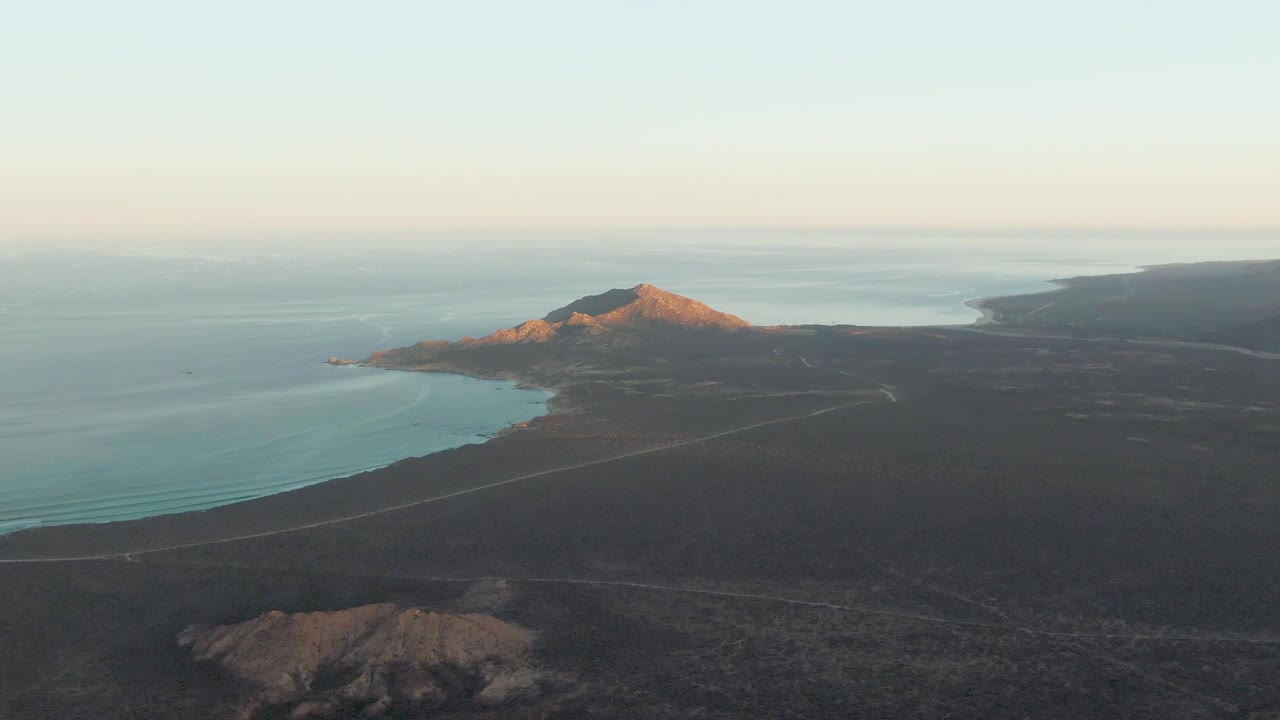 Aerial view over Cabo Pulmo, Baja California Sur showing a pale coastal road through dark terrain beside blue ocean, with a sunlit mountain ridge in the distance and no visible people.