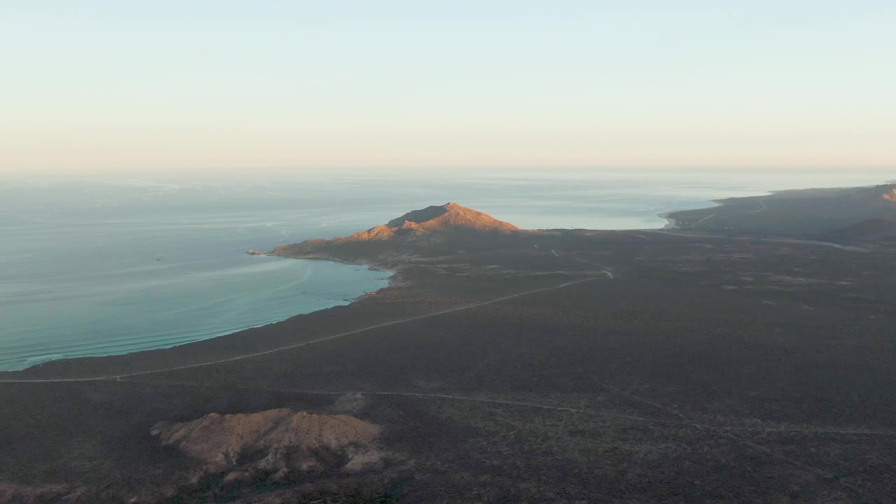 Wide aerial frame over Cabo Pulmo with the road running across dark coastal terrain, blue water curving along the shore, a rocky mountain ridge catching warm light, and no visible people.