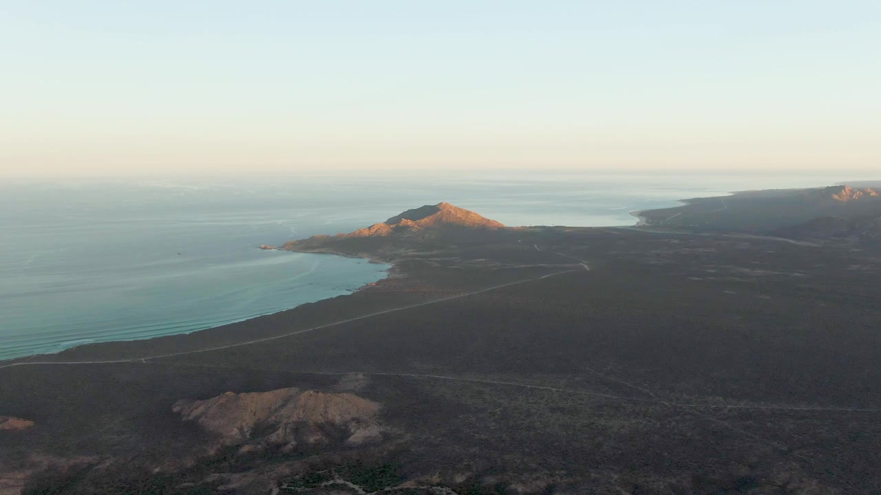 Cabo Pulmo aerial frame with dark foreground land, a thin coastal road, turquoise water, a low sunlit mountain ridge near the shore, distant headlands, and no visible people.