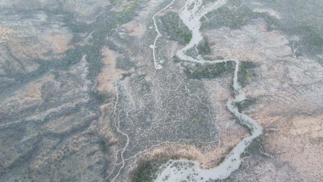 Top-down drone view of a dry channel bending through muted tan and gray arid land in Baja California Sur, with small green shrub clusters and no visible people.