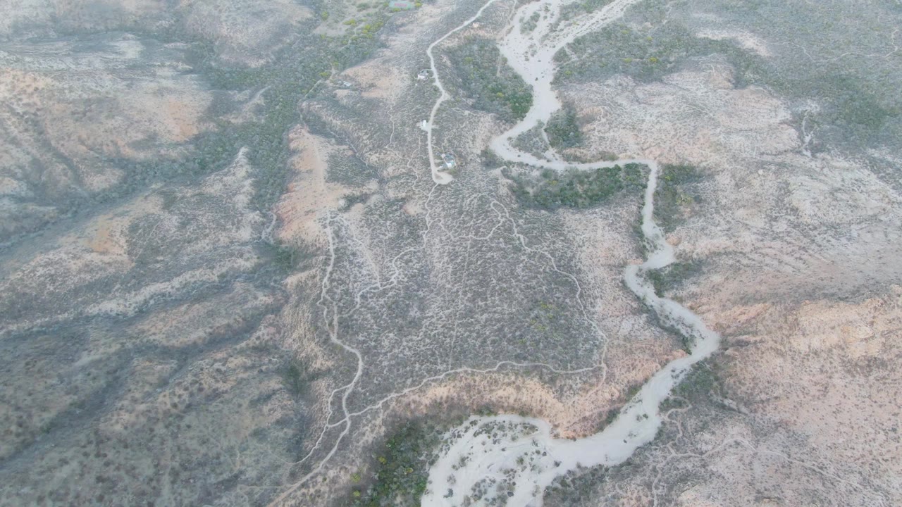 Overhead Cabo Pulmo aerial with the pale river channel bending through a broad gray wash, tan slopes, small green shrubs, and no visible people.