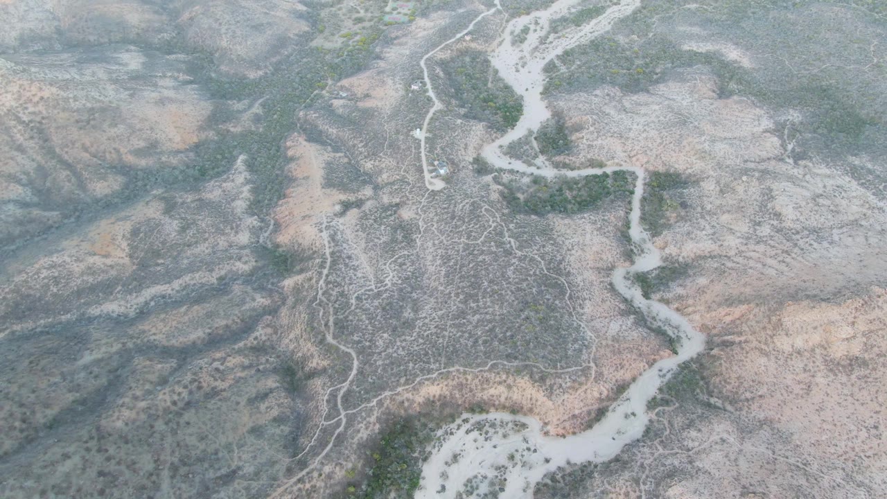 Cabo Pulmo overhead frame showing a braided pale wash running from the upper edge toward the center of gray-brown desert terrain, with green patches and no visible people.