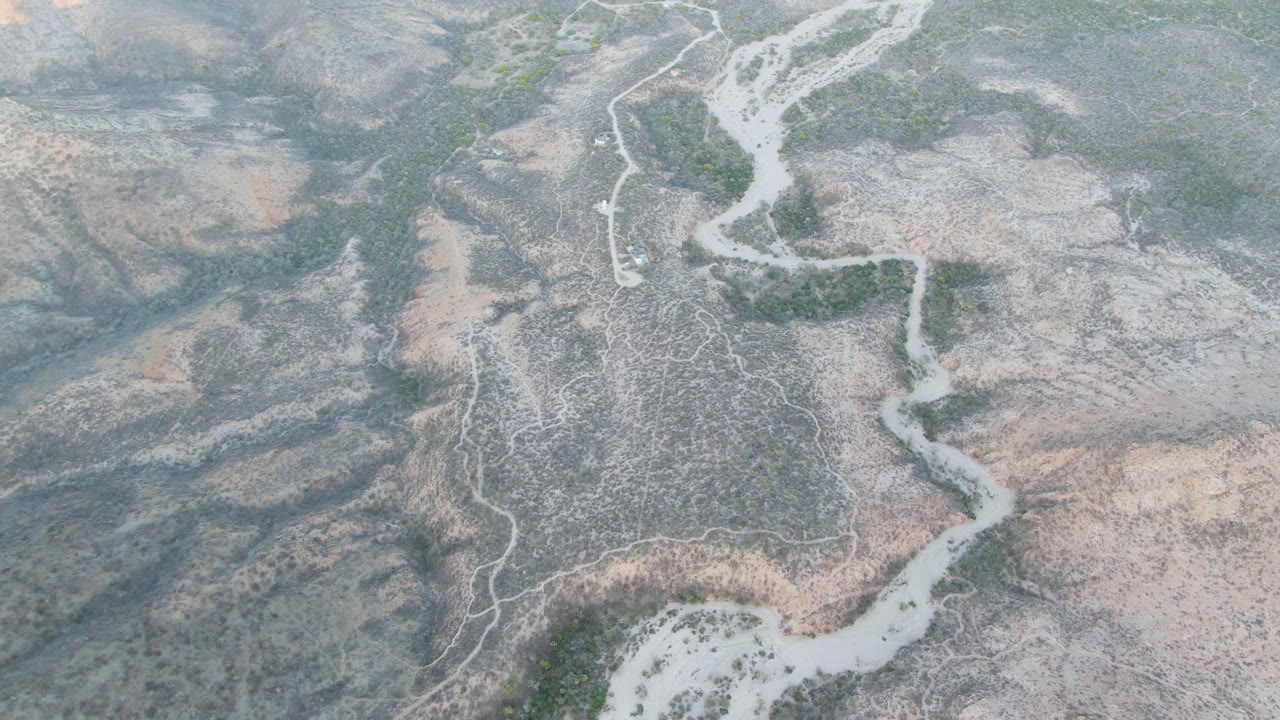 Later overhead frame from Cabo Pulmo with the winding pale channel crossing a darker gray wash area, bordered by tan arid slopes, green vegetation clusters, and no visible people.