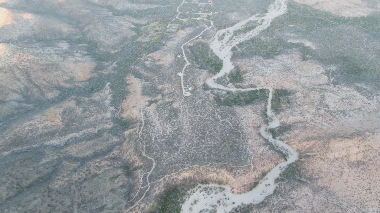 Aerial frame over Cabo Pulmo, Baja California Sur, showing a pale winding riverbed crossing rocky hills with scattered green vegetation and no visible people.