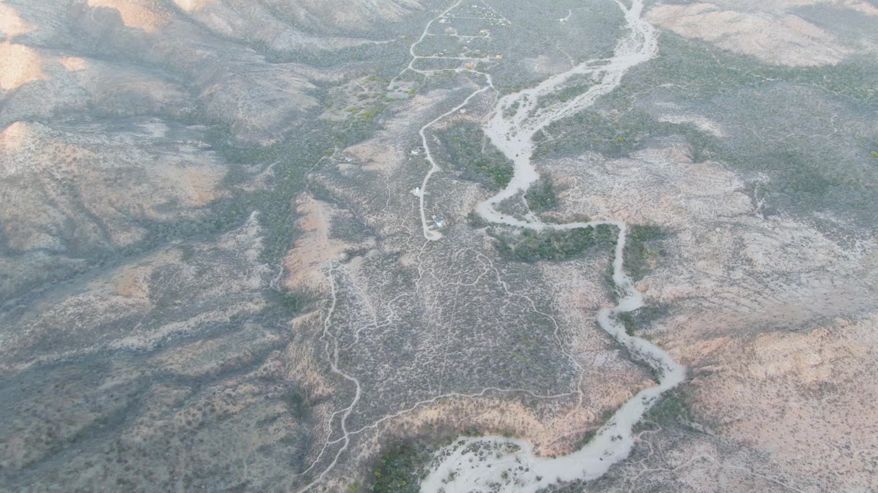 High drone-style frame over Cabo Pulmo, Baja California Sur, showing the pale river channel curving between arid hills, green pockets, and no visible people.