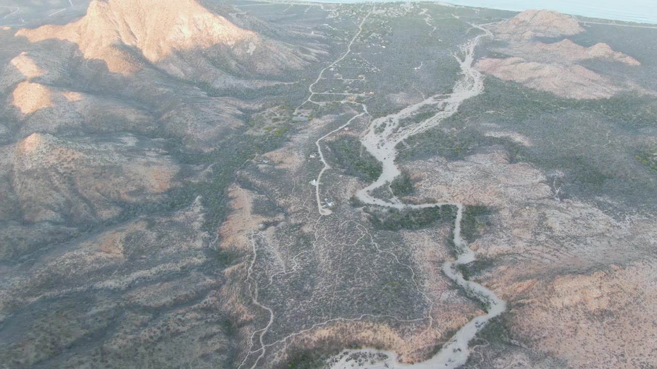 Aerial frame over Cabo Pulmo terrain with the pale wash and road crossing the center, darker scrub on the slopes, sunlit mountain faces in the distance, and no visible people.