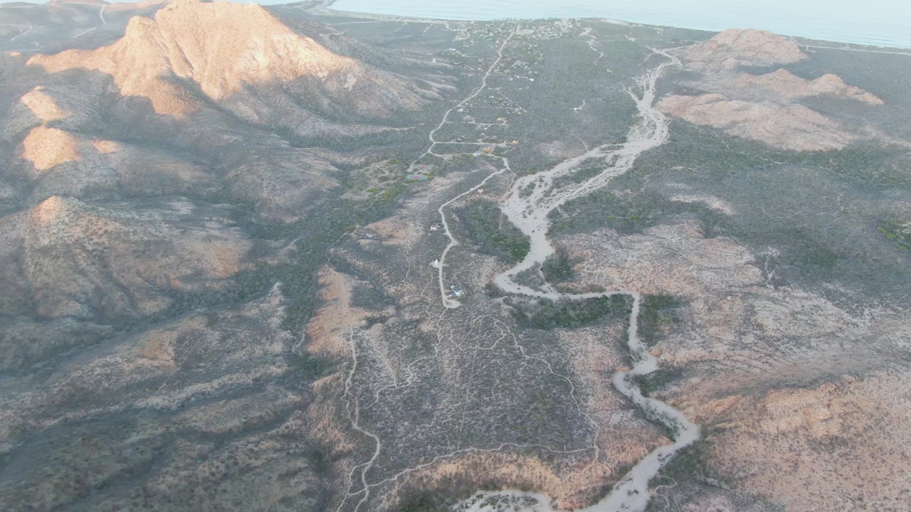 Mid-clip aerial frame in Cabo Pulmo showing a branching dry riverbed, a winding dirt route, muted gray and tan slopes, background mountains, and no visible people.