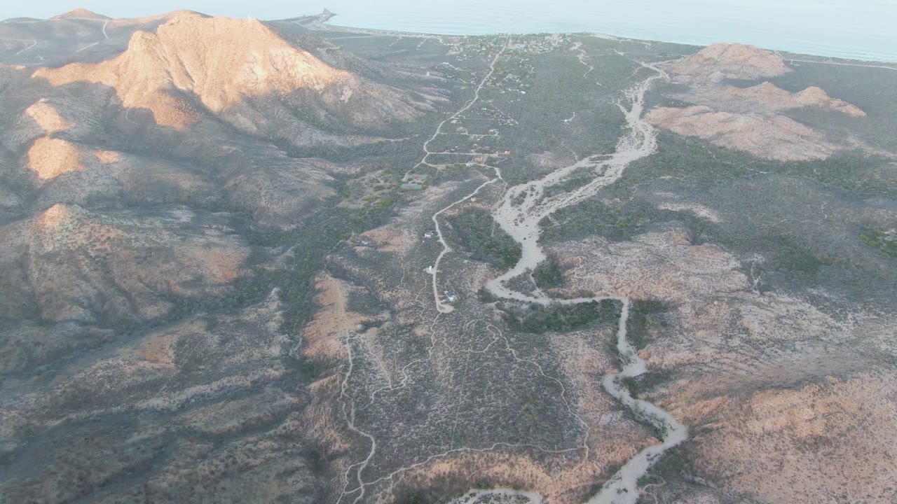 Later aerial frame over Cabo Pulmo with the road and wash shifted across the center, broad rocky flats in the foreground, mountain ridges beyond, and no visible people.