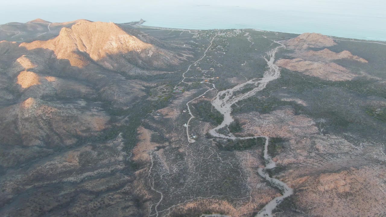 Aerial frame over Cabo Pulmo arid hills, with a winding pale road crossing scrub terrain below a sunlit mountain ridge and no visible people.
