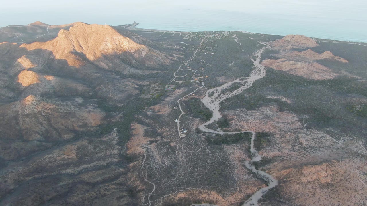 Aerial view near Cabo Pulmo showing the dirt road bending through darker scrubland, with bright light on the left ridge and no visible people.