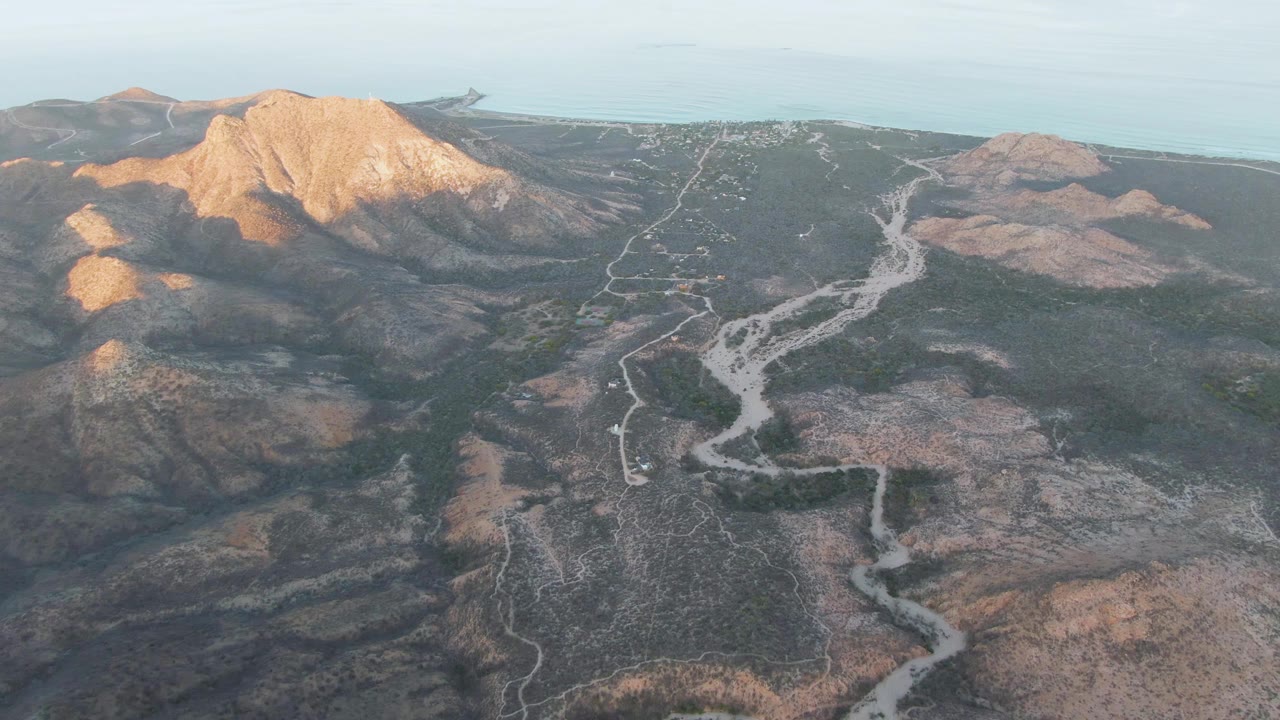 Cabo Pulmo aerial frame with the road running from the lower terrain toward the center, a bright mountain shoulder at left, and no visible people.