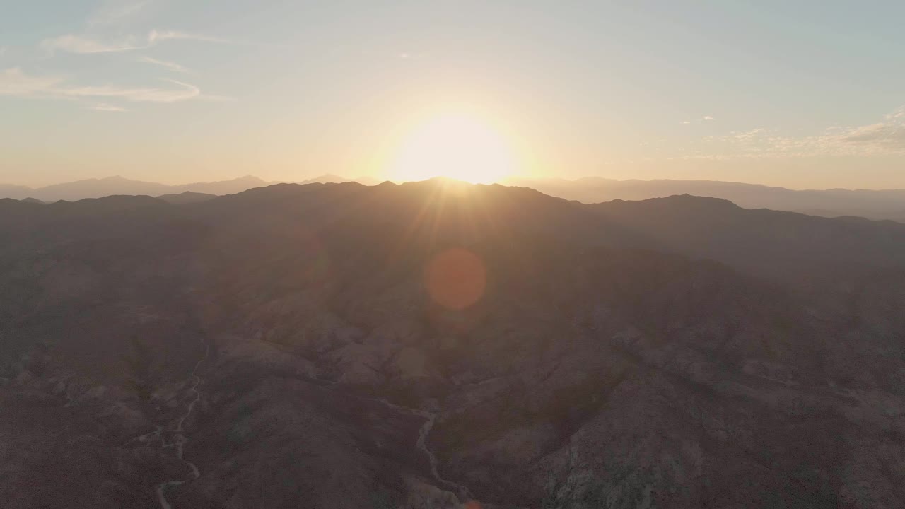 At 0:00, the sun sits above layered arid mountains near Cabo Pulmo, Baja California Sur, with a small orange lens flare and no visible people.