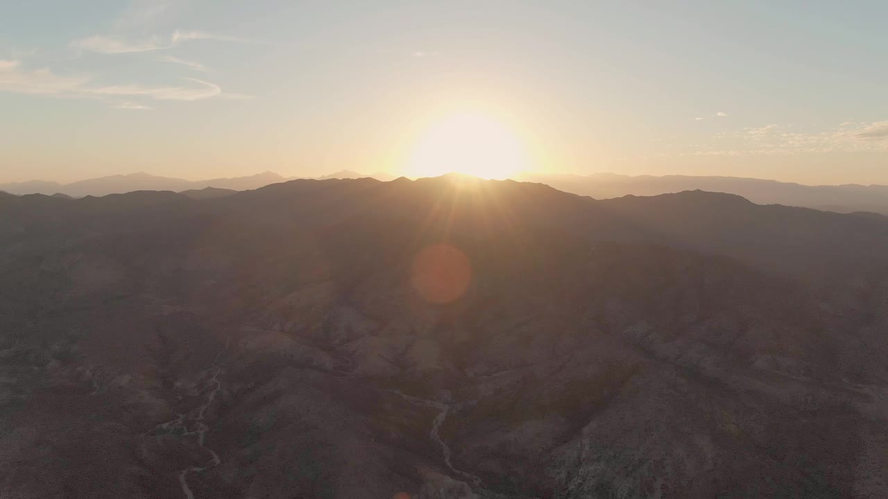 At 0:02, the low sun shines across rugged dry mountains near Cabo Pulmo, with a warm flare over the shadowed valley and no visible people.