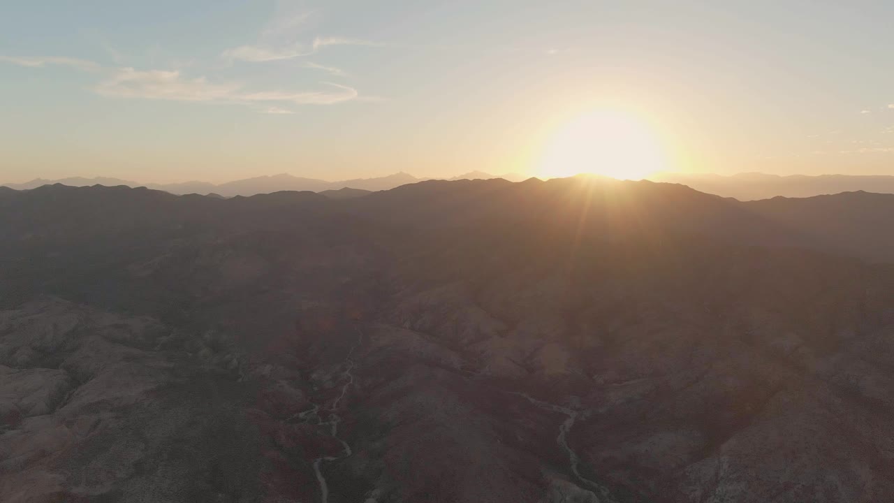 Wide aerial frame of the sun touching the mountain horizon above Cabo Pulmo canyon ridges, with a winding valley channel below and no visible people.