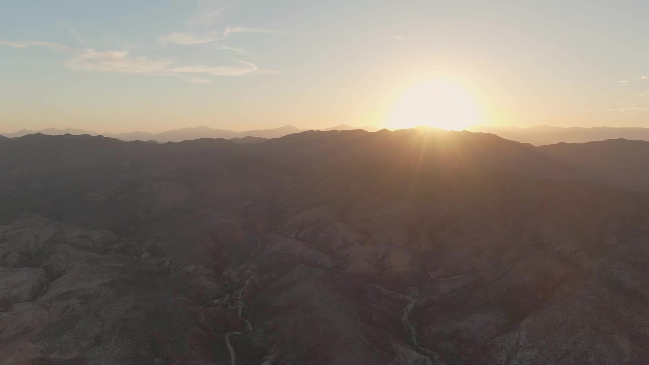 Wide Cabo Pulmo aerial composition with red-brown ridges, a faint winding stream or wash in the valley, and the sun flaring over the mountains with no visible people.