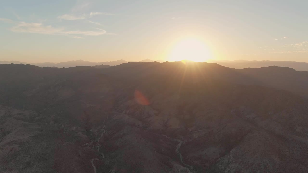 Low sun breaks over mountain ridges above a shadowed Cabo Pulmo valley, with a pale winding river channel below and no visible people.