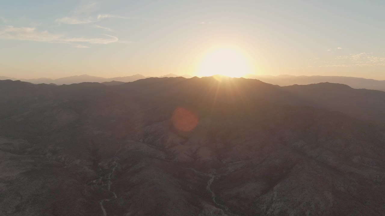 Golden light spreads from the sun over the Cabo Pulmo mountain horizon, revealing ridged terrain and a faint riverbed track in the valley with no visible people.