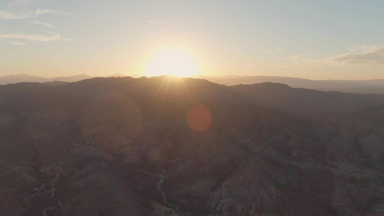 At 0:01, the sun flares over layered Cabo Pulmo mountain silhouettes, with rugged brown terrain and a winding channel visible through the dark valley, no visible people.
