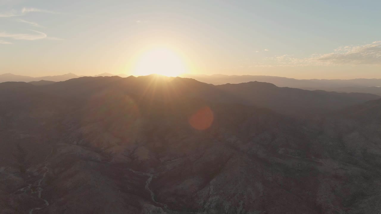 At 0:02, the aerial view holds on the Cabo Pulmo valley as the sun glows above the ridge, with distant mountain bands, dark rocky slopes, and a faint winding watercourse or wash, no visible people.