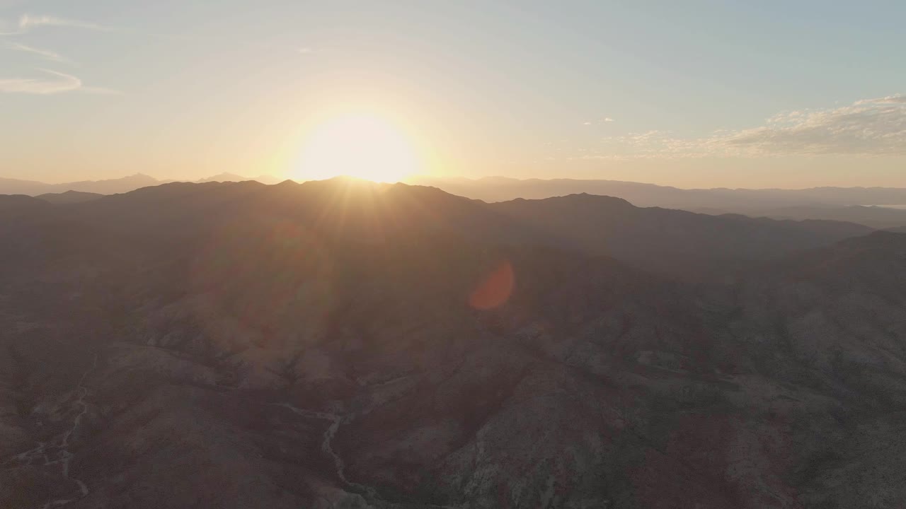 At 0:03, the sun is brighter near the upper left of the frame over Cabo Pulmo's arid mountain ridges, with a dark valley floor and winding channel beneath, no visible people.