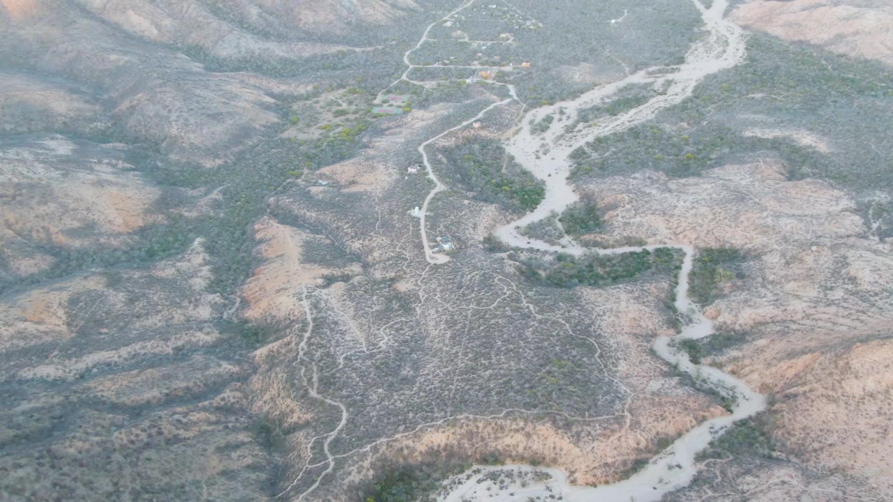 Aerial frame from Cabo Pulmo, Baja California Sur showing a pale riverbed running through a dry valley floor, surrounded by low hills and scattered shrubs with no visible people.