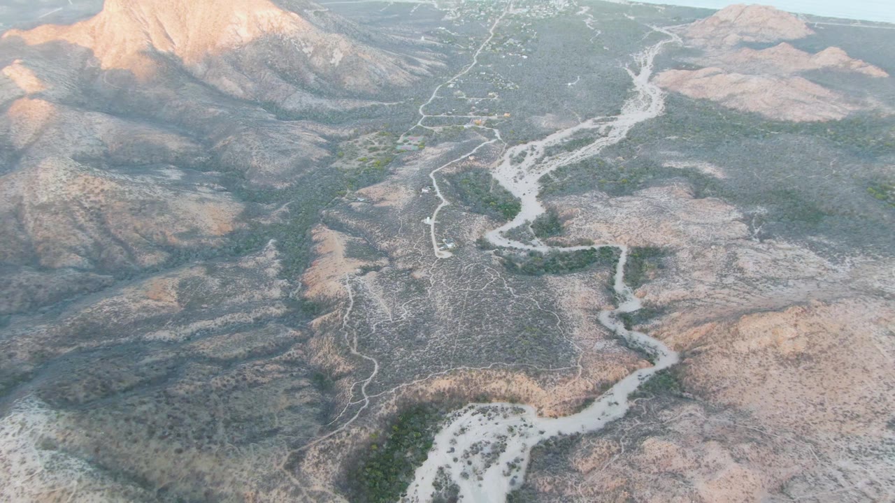 Drone-height view in Cabo Pulmo showing the dry wash bending through rocky brown terrain, with green brush patches and sunlit mountain slopes in the background, no visible people.