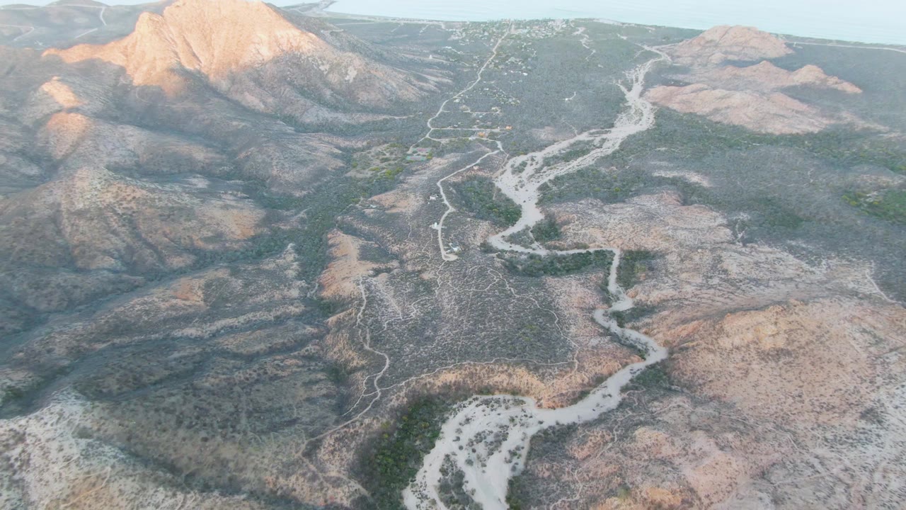 High aerial frame over Cabo Pulmo with the pale riverbed threading from the lower valley toward distant arid mountains, bordered by scrub vegetation and no visible people.