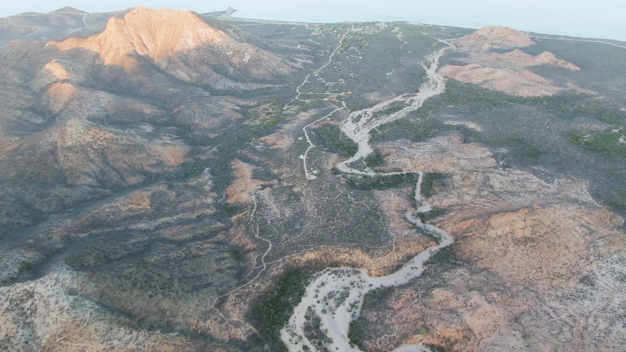 Aerial view in Cabo Pulmo with a sunlit mountain mass at upper left, a winding dry riverbed through the center, scattered green vegetation, and no visible people.