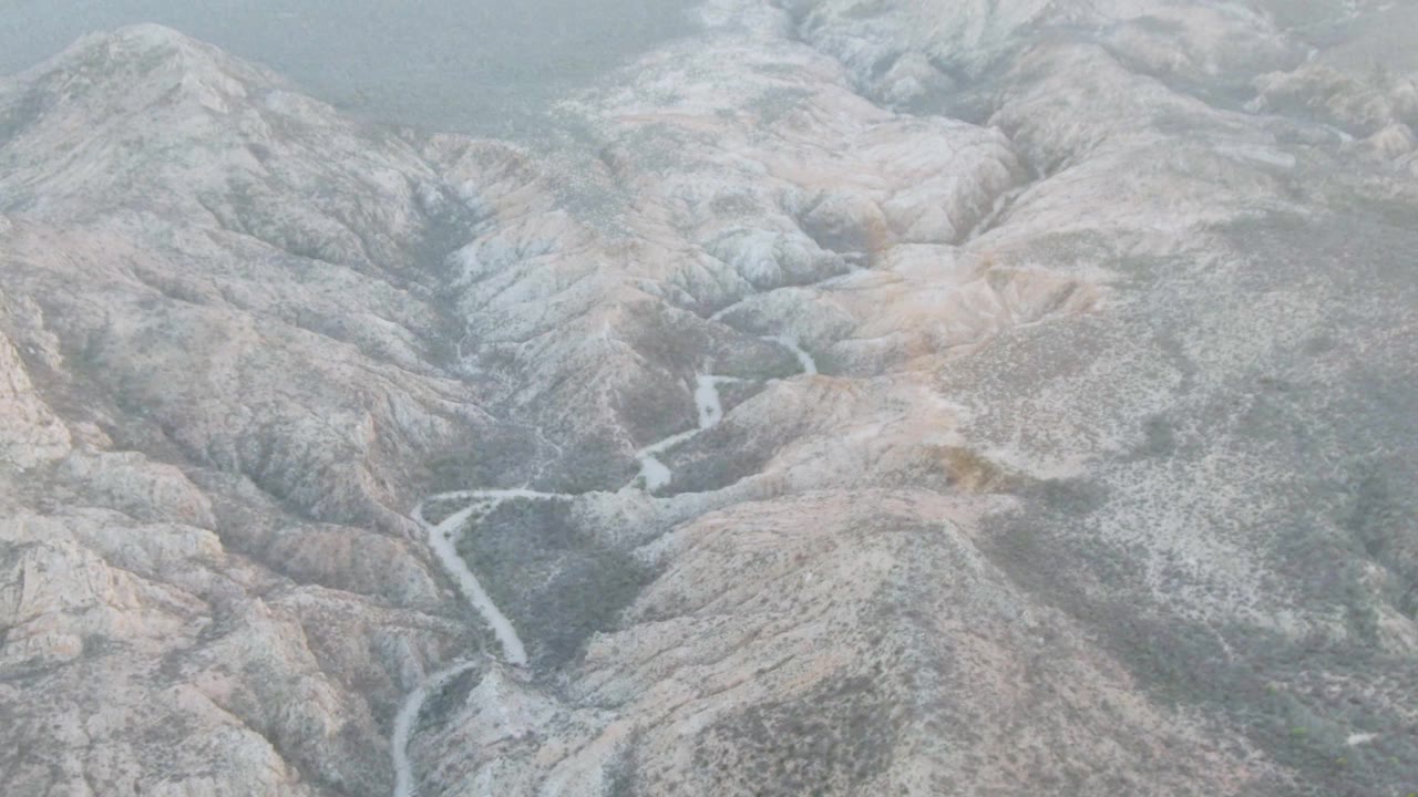 Aerial view over a rocky mountain valley near Cabo Pulmo, Baja California Sur, with a faint rainbow in hazy light, a winding stream below, and no visible people.