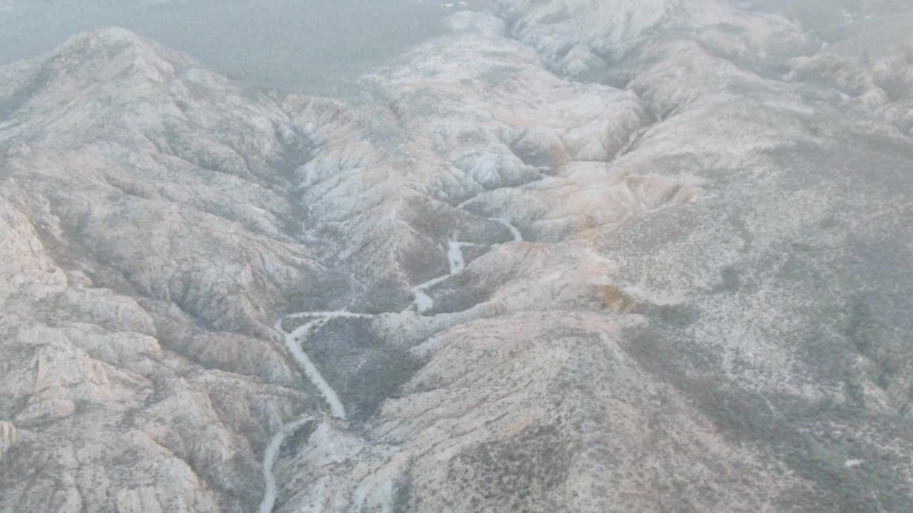 Aerial frame over the Cabo Pulmo area mountain terrain, with pale ridges, a narrow stream crossing the valley floor, softened haze, and no visible people.
