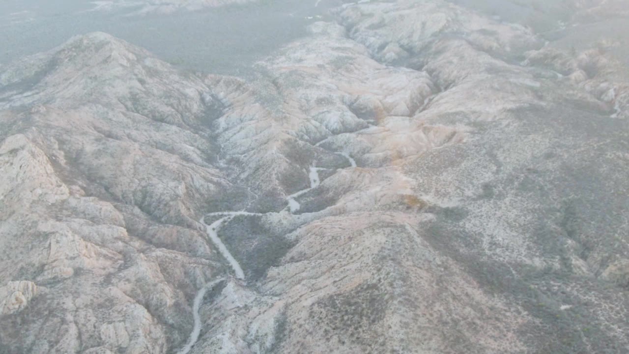 High aerial view of rocky mountains in Baja California Sur, with a pale winding stream in the midground, subdued cloud-filtered light, and no visible people.