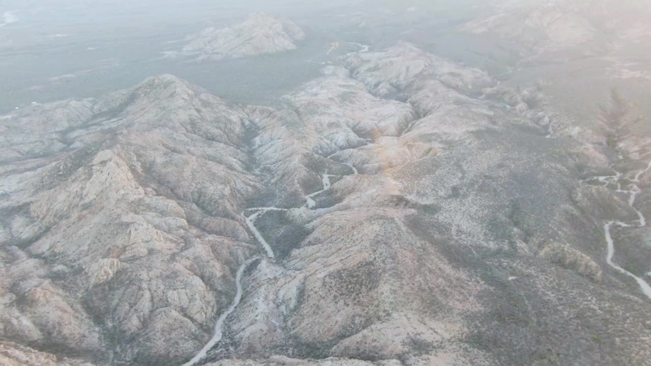 Aerial view over Cabo Pulmo rocky mountain ridges with pale winding channels crossing the valley floor, hazy water near the horizon, and no visible people.