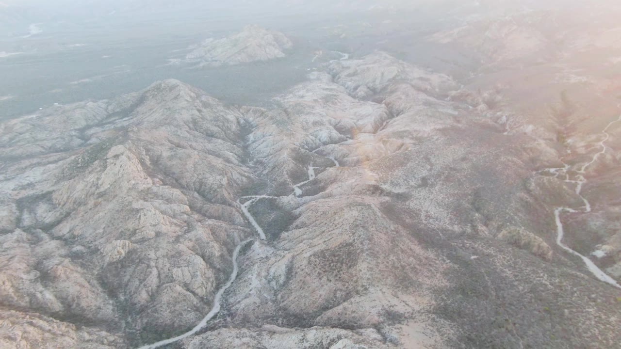 Wide aerial frame in Cabo Pulmo showing gray-brown ridges, a pale winding riverbed or drainage through the center, hazy blue water at top, and no visible people.