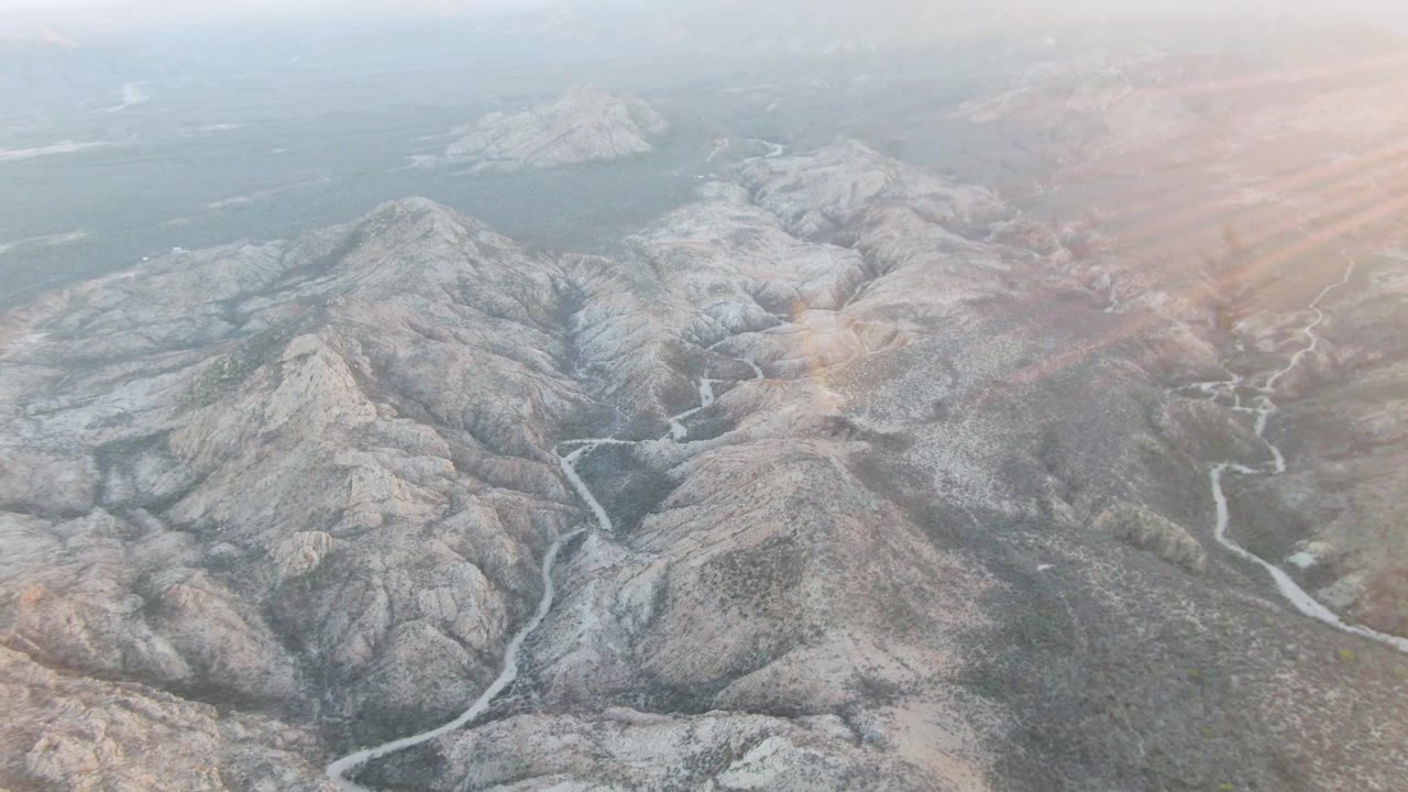 Aerial view over Cabo Pulmo mountain terrain with branching pale channels in the valley, muted rocky slopes, warm haze at the upper right, and no visible people.