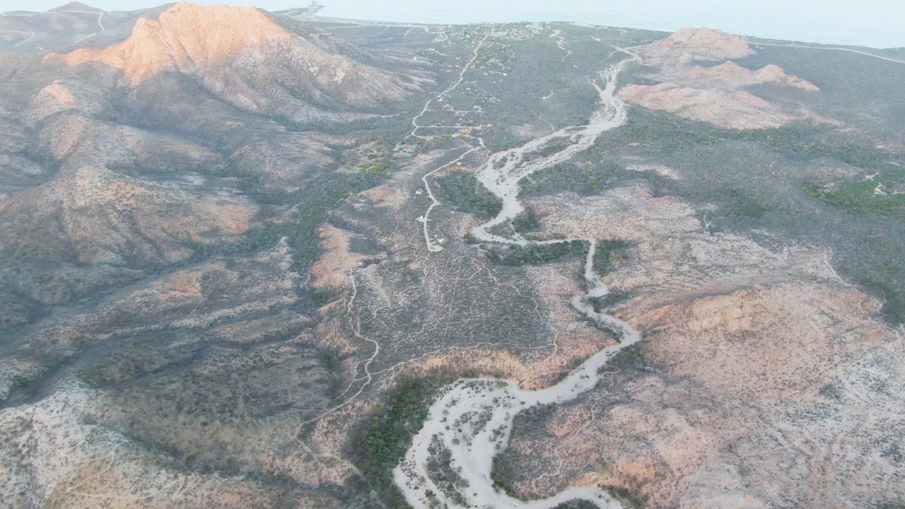 Aerial view over Cabo Pulmo, Baja California Sur, showing a pale winding riverbed crossing an arid valley below rocky mountains, with no visible people.
