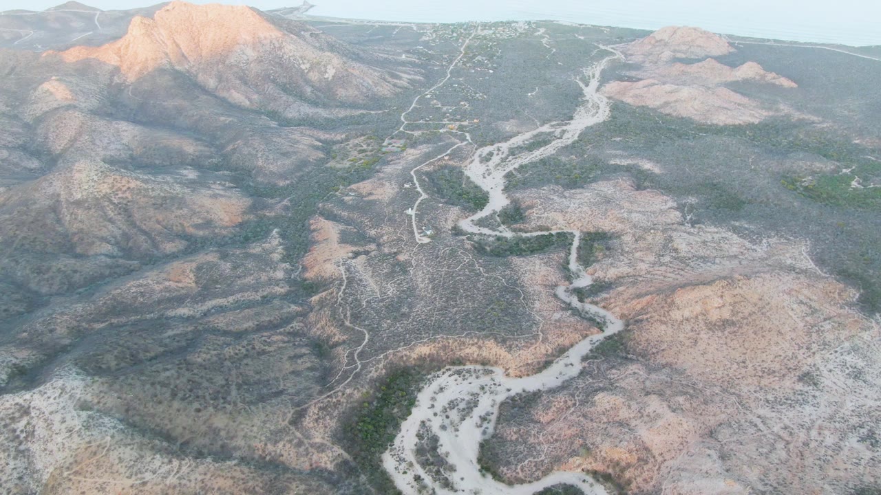 High aerial frame in Cabo Pulmo, Baja California Sur, with the branching pale wash running through dry hills toward sunlit mountains, and no visible people.