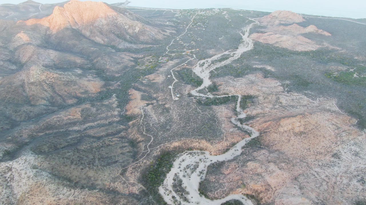 Aerial landscape over Cabo Pulmo, Baja California Sur, showing the winding wash and green patches set against dry earth-toned hills, with no visible people.