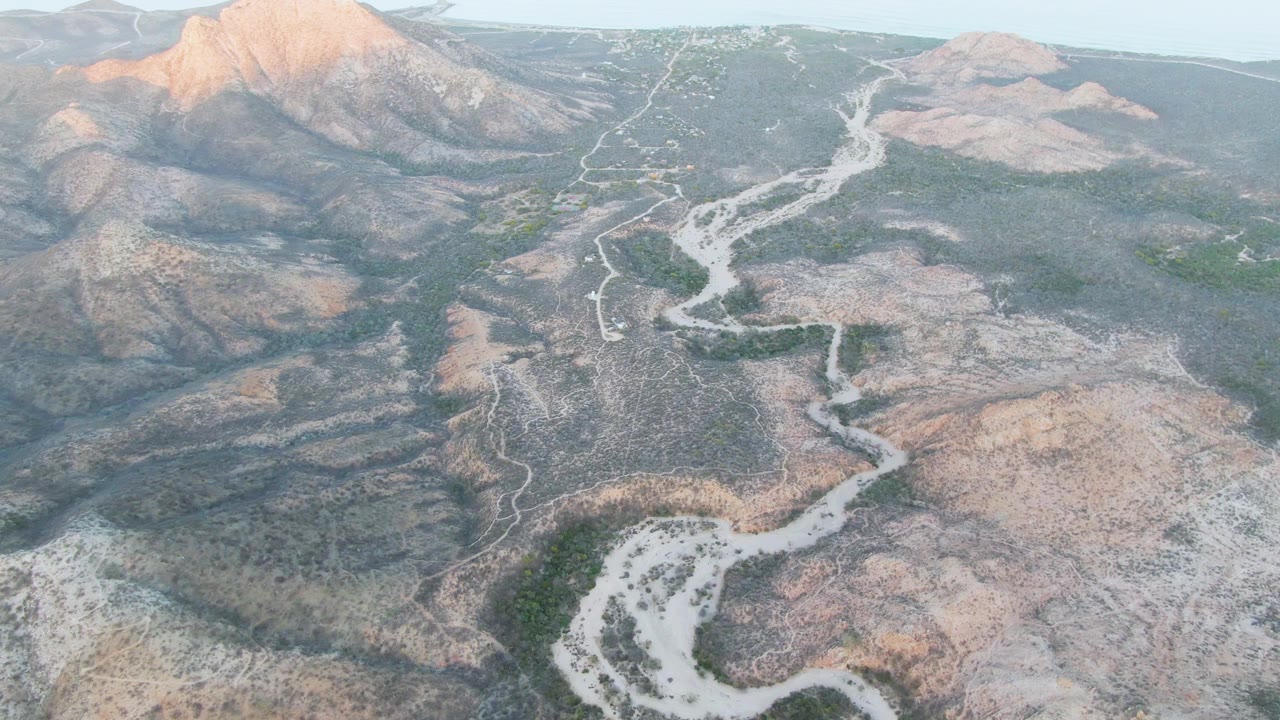 High oblique aerial frame from Cabo Pulmo showing the winding pale wash nearer the center, rocky tan and gray slopes, scattered green patches, and no visible people.