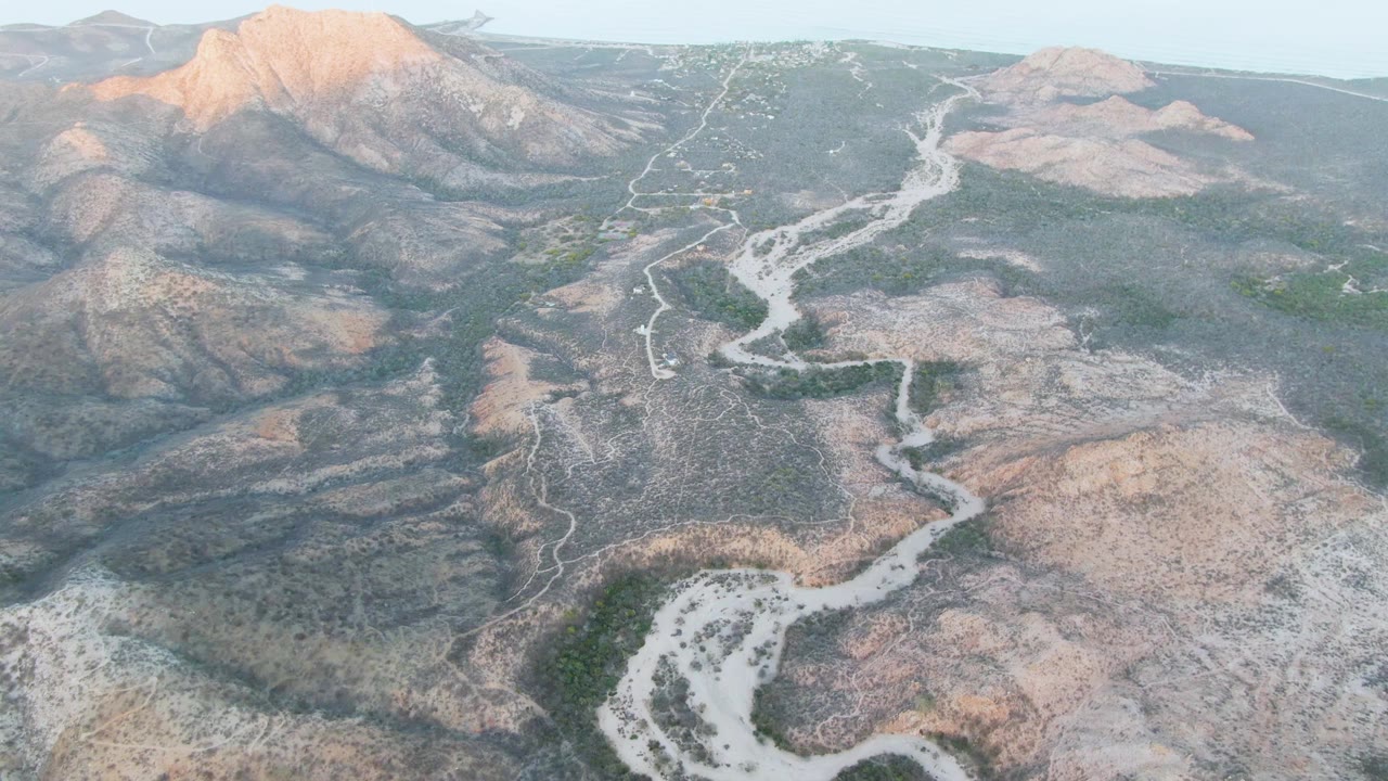Aerial view in Cabo Pulmo with the foreground wash crossing the lower frame, broad rocky hills beyond it, warm light on the mountain face, and no visible people.