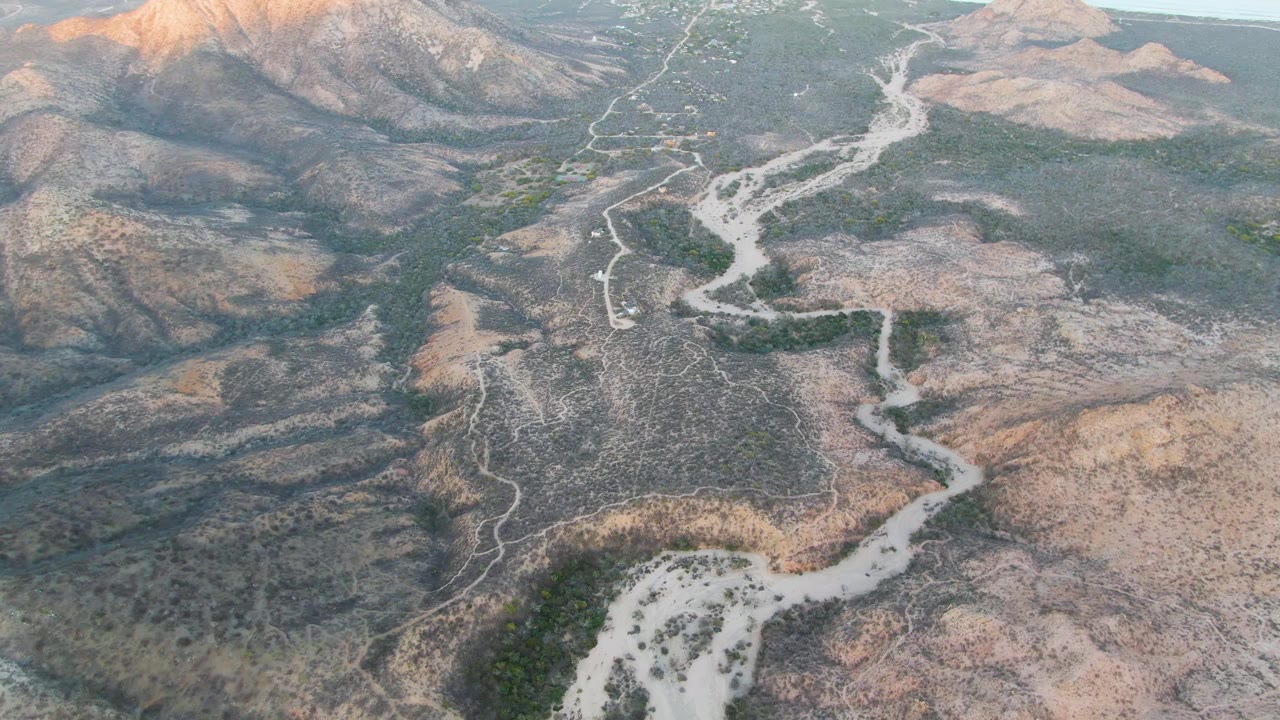 Aerial frame over Cabo Pulmo dry terrain, with the dirt road bending through a broad wash, sparse green vegetation, warm upper slopes, and no visible people.