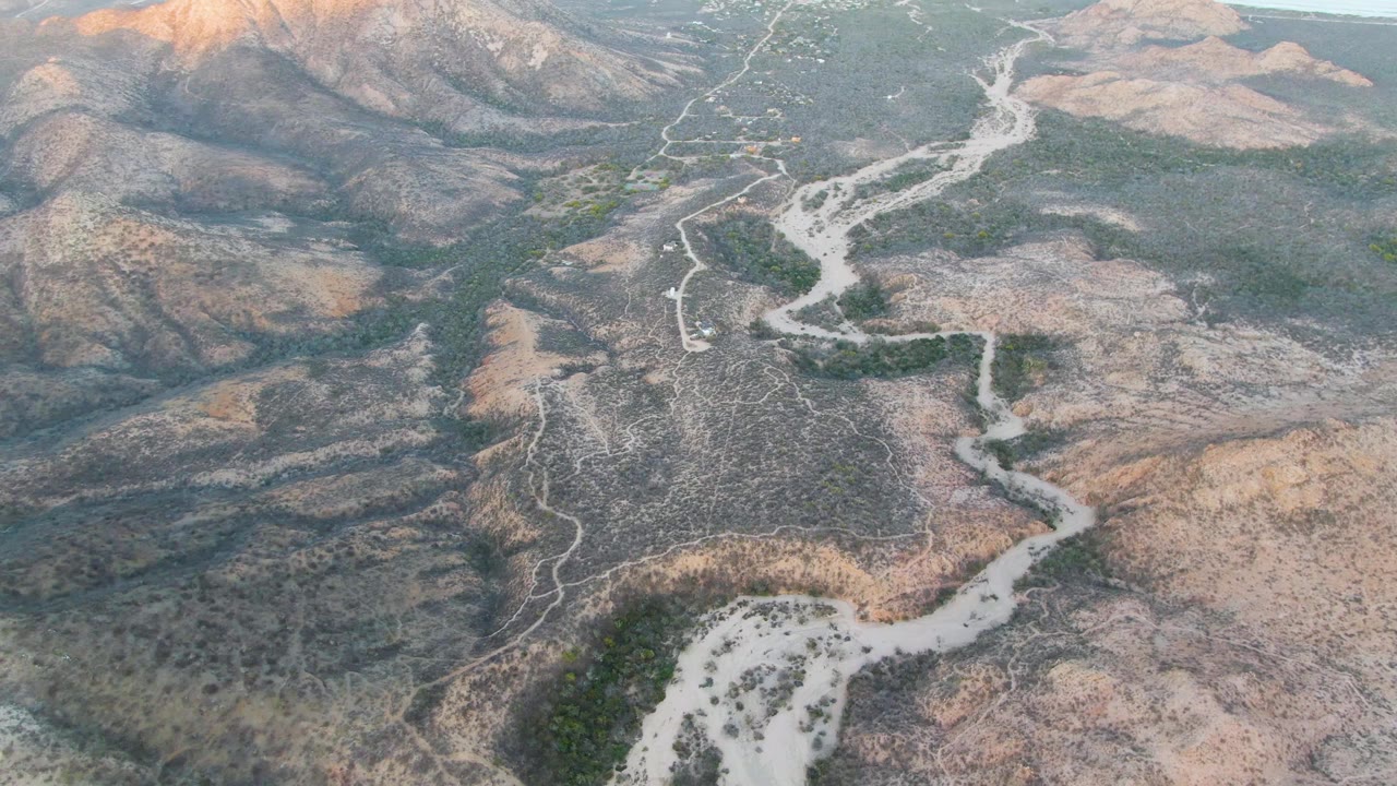 Aerial view in Cabo Pulmo showing the road and pale wash lower in frame, mottled brown terrain across the valley, green shrub clusters, and no visible people.