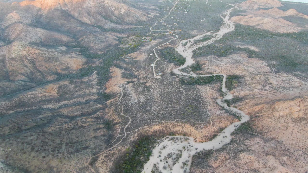 Aerial frame over Cabo Pulmo dry hills shows a pale winding dirt road cutting through brown ridges and green valley patches, with no visible people.