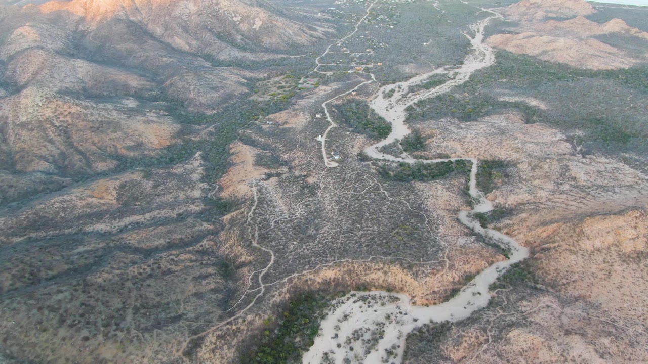 Drone-like view in Cabo Pulmo shows the pale road bending across rugged brown hills, with darker green vegetation following low valley areas and no visible people.