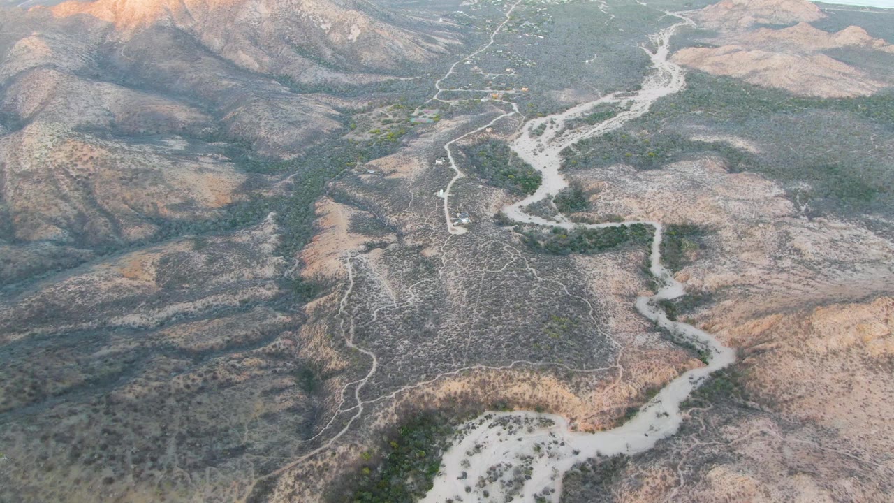 Elevated Cabo Pulmo frame shows a winding mountain road running from the lower frame toward dry ridges and muted green valleys, with no visible people.