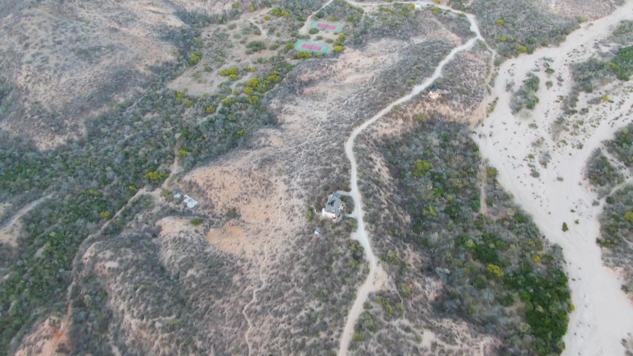High aerial view near Cabo Pulmo, Baja California Sur, showing dry hills, a pale winding dirt road, scattered green shrubs, a sandy wash, and no visible people.