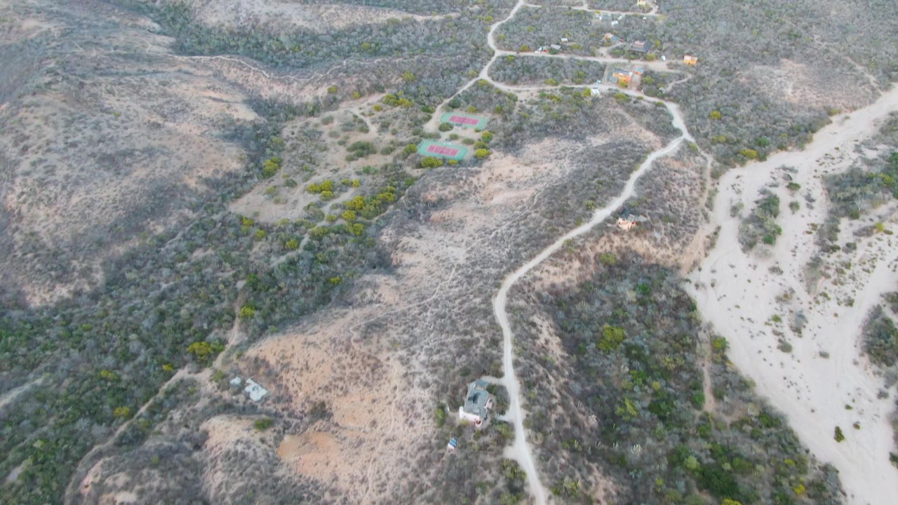 Wide drone view near Cabo Pulmo showing an arid hillside road, a pale sandy wash through the valley floor, small structures, scattered shrubs, and no visible people.