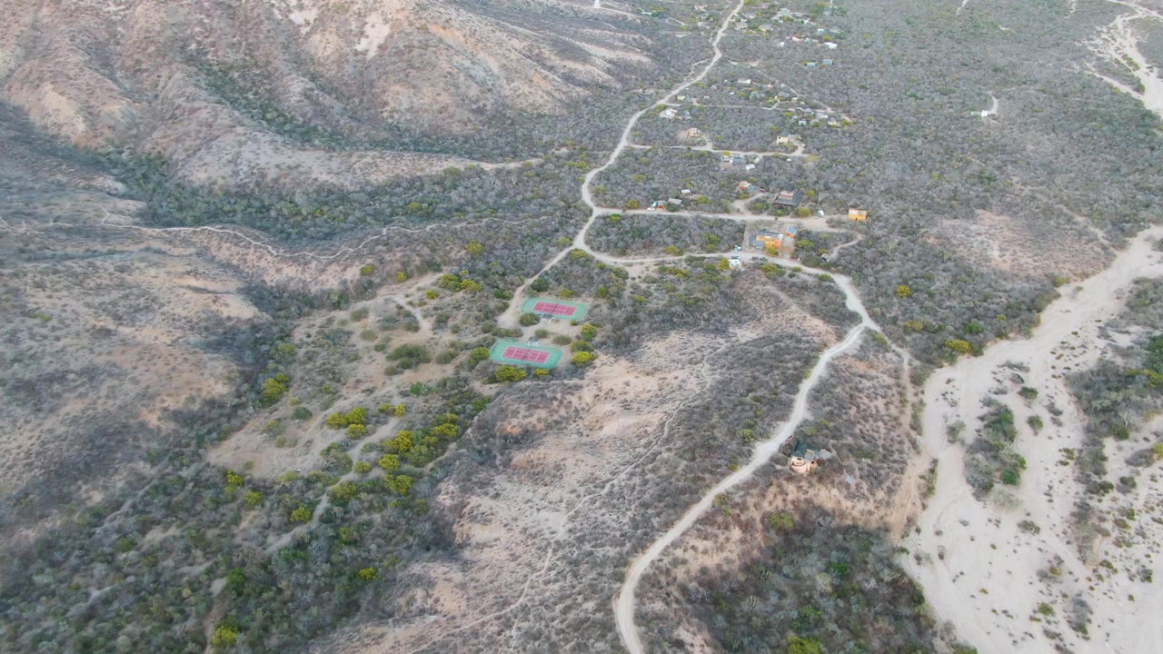 Aerial frame over Cabo Pulmo showing dry scrub hills, a pale wash, winding dirt roads, small buildings, and no visible people at the start of the clip.